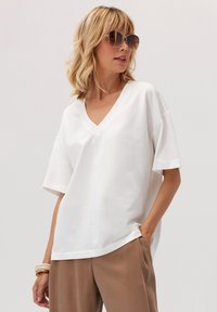 Biała bawełniana koszulka V-neck z krótkim rękawem, luźny krój, w połączeniu z jasnobrązowymi spodniami. Modelka w oversize'owych okularach przeciwsłonecznych.