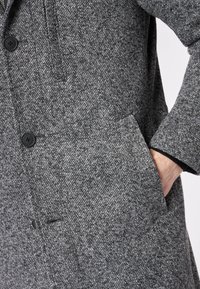 Grau strukturierter Mantel mit feinem Gewebe, zwei vorderen Taschen und einem schwarzen Knopfdetail. Der Stoff wirkt dick und warm.