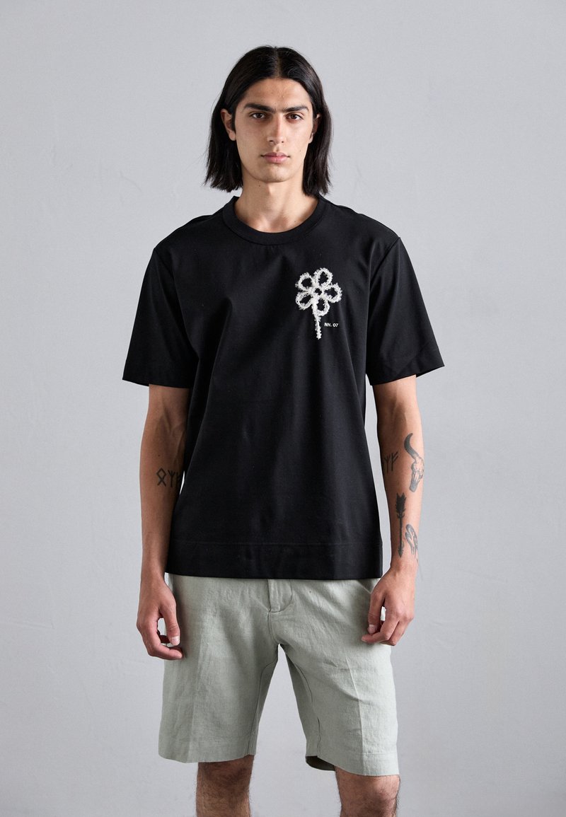 Sort bomulds t-shirt med et struktureret hvidt blomstermotiv på brystet, parret med lysegrønne linnedshorts. Korte ærmer og rund hals.