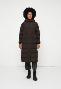 Lungo cappotto piumino nero con cappuccio, caratterizzato da un design trapuntato, zip anteriore a tutta lunghezza e maniche larghe. Indossato sopra pantaloni grigi e stivali neri.