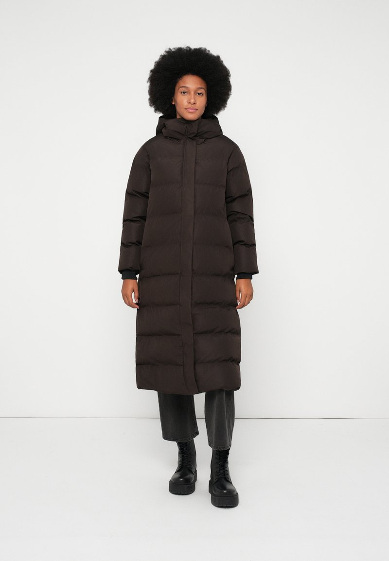 Lungo cappotto piumino nero con cappuccio, caratterizzato da un design trapuntato, zip anteriore a tutta lunghezza e maniche larghe. Indossato sopra pantaloni grigi e stivali neri.