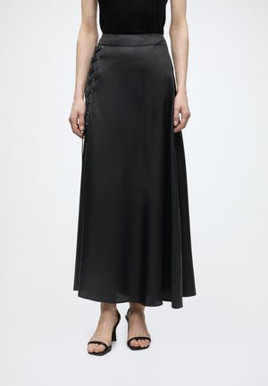 Čierna saténová sukňa po členky s prešitou šnurovacou detailom na ľavej strane, nosená s čiernym tielkom bez rukávov a čiernymi otvorenými sandálmi na podpätku.