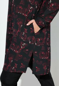 Abito lungo nero a maniche lunghe con un motivo geometrico rosso, dotato di tasca e fessure laterali. Realizzato in tessuto morbido e testurizzato.
