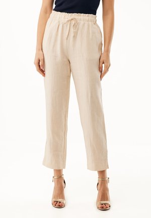 Pantalon beige en lin avec cordon de serrage, porté avec un top noir sans manches et des sandales beige à talons hauts sur fond blanc.