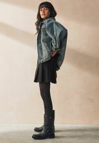 Giacca in denim con maniche larghe, abito nero, collant opachi a motivi e stivaletti in pelle nera con fibbie. Colori e texture solidi.
