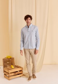 Lichtblauwe overhemd met bloemenpatroon, gecombineerd met beige broek en bruine schoenen, staande naast houten kratten en citroenen.