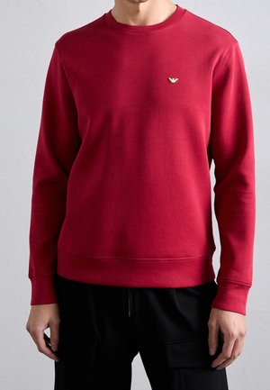 Mand iført ensfarvet rød langærmet sweatshirt med lille gyldent logo på brystet, kombineret med sorte bukser mod ensfarvet baggrund.