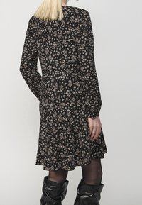 Femme aux cheveux blonds portant une robe longue à manches longues à motifs floraux noirs, des collants noirs et des bottes en cuir noires décontractées, vue de dos.