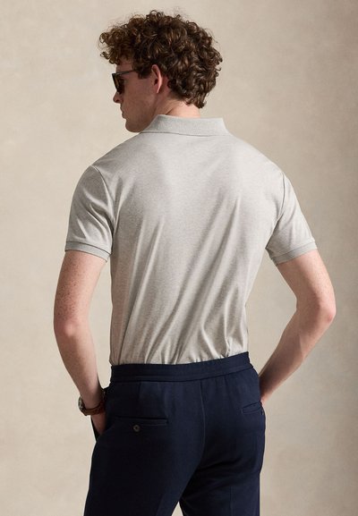 Homme aux cheveux bouclés portant des lunettes de soleil, polo gris clair à manches courtes rentré dans un pantalon bleu marine foncé, debout les mains dans les poches.