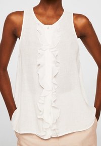 Blusa sin mangas blanca con detalle de volantes en la parte delantera, escote redondo y una tela ligera. Presenta un cierre de botón en la parte superior.