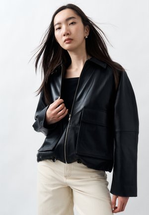 Junge Frau mit langen dunklen Haaren, die eine aufgeknöpfte schwarze Lederjacke und hellbeige Hosen vor einem schlichten hellen Hintergrund trägt.