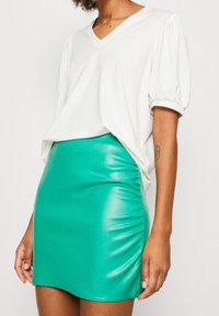 Biała koszulka typu V-neck z bufkami, zestawiona z mini spódnicą w kolorze turkusowym o błyszczącej powierzchni, sięgającą do kolan, z detalem marszczonym po boku.