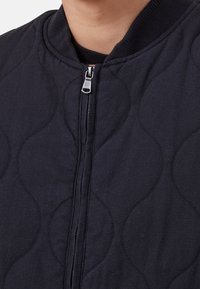 Czarna pikowana kurtka z zamkiem błyskawicznym z przodu, prążkowanym kołnierzem oraz teksturowanymi falistymi wzorami na tkaninie dla dodatkowej głębi.