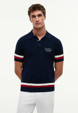 Młody mężczyzna w granatowej koszulce polo z czerwono-białymi paskami na rękawach i dole oraz białych spodniach, stojący z rękami za plecami.