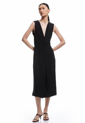 Mujer de pie con las manos en las caderas, vestida con un vestido midi negro sin mangas con escote en V profundo y sandalias negras de tacón con tiras.