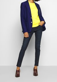 Námořnický blejzr přes zářivě žlutý top, tmavé úzké džíny a burgundské boty s texturovanými akcenty. Blejzr má dvě knoflíky a kapsy.