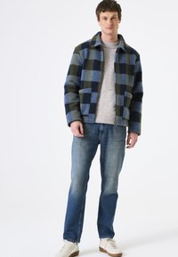 Mann in einer blau-grünen karierten Jacke, leicht gesprenkeltem Sweater, blauen verwaschenen Jeans und weißen Sneakers, der vor einem schlichten weißen Hintergrund steht.