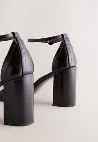 Paire de chaussures en cuir marron foncé à talons blocs avec brides à la cheville sur un fond neutre.