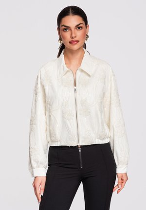 Kvinde iført en hvid, blomstret, tekstureret jakke med lynlås og sorte bukser, stående foran en ensfarvet lys grå baggrund.