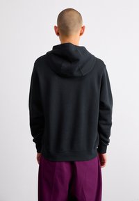 Felpa nera con cappuccio, con polsini e orlo a costine, realizzata in tessuto morbido. La vista posteriore non mostra loghi o motivi visibili. Pantaloni viola visibili sotto.