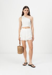 Fehér, egy vállra szabott crop top, fehér, fodros sorttal párosítva. A modell szőtt kosár táskát tart, fekete papucsot visel. Egyszerű, könnyed kialakítás.