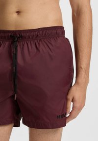 Bordowe kąpielówki wykonane z lekkiego materiału, z elastycznym paskiem w talii, sznurkiem do regulacji, bocznymi kieszeniami oraz czarnym logo.