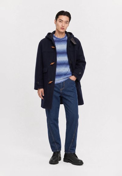 Junger Mann trägt einen marineblauen Toggle-Mantel, blauen gemusterten Pullover, blaue Jeans und schwarze Schnürstiefel, steht mit einer Hand in der Tasche.