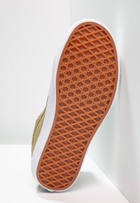 Braune Gummisohle mit Rauten- und Sechseckprofil auf beigem Sneaker, der vor weißen Wänden positioniert ist.