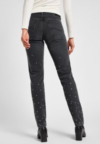 Jeans de ganga pretos com um corte justo, apresentando ornamentações brancas espalhadas e bolsos traseiros. O tecido tem uma textura suave.