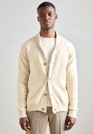 Cárdigan de punto beige con cuello de chal, con un patrón texturizado y botones dorados, llevado sobre una camisa de color claro.
