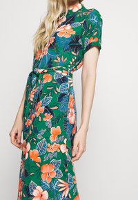Sukienka midi w kwiaty ma zieloną bazę z pomarańczowymi i niebieskimi wzorami kwiatowymi, krótkie rękawy, pasek w talii oraz luźny krój.