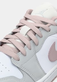 Baskets en cuir blanc avec des accents en daim gris et des lacets rose doux. Des surfaces texturées et de subtils détails de branding sont visibles.