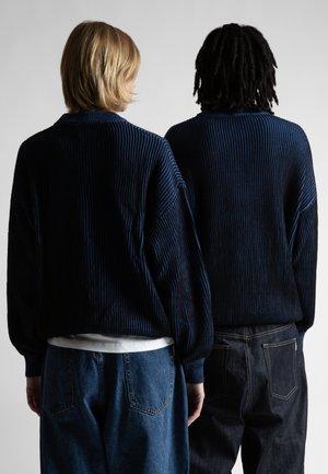 To personer står side om side, vendt væk, iført matchende mørkeblå ribbede trøjer og denimjeans mod en ensfarvet baggrund.