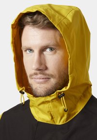 Chaqueta amarilla con capucha y cuerpo negro. Presenta un tejido texturizado, cordones ajustables y malla de ventilación en el interior de la capucha.