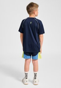 Marint kortärmad sporttröja med reflekterande detaljer, tillsammans med blå och neongröna shorts. Skorna är vita och strumporna är mönstrade.