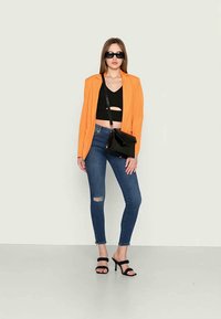 Blazer laranja sobre um top preto curto, combinado com jeans azuis desgastados e sandálias pretas de salto. Os acessórios incluem uma pequena bolsa preta.