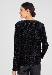 Suéter de terciopelo negro con rayas verticales de lentejuelas, mangas largas y un amplio escote redondo. Combinado con pantalones de cuero negros.