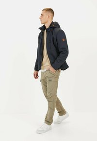 Dunkelblaues Softshell-Jacke mit Kapuze, mit Reißverschlusstaschen; getragen wird ein beiger Pullover und khakifarbene Cargo-Hosen, ergänzt durch weiße Sneakers.