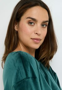 Top in velluto verde acqua con una consistenza morbida e scollatura ampia, caratterizzato da leggere plissettature sulle spalle. Il modello indossa piccoli orecchini a cerchio dorati.