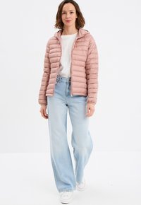 Giacca puffer rosa chiaro con baffi orizzontali, chiusura con zip e cappuccio integrato, abbinata a jeans azzurri a vita alta e sneaker bianche.