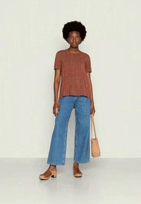 Top curto de manga curta com padrão castanho, jeans largos azuis, sandálias de dedo em tom bege e bolsa de palha. Modelo posa contra um fundo neutro.