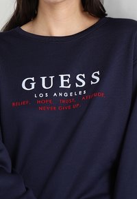 Námořnická modrá mikina se kulatým výstřihem, s bílým a červeným textem: "GUESS LOS ANGELES" a motivačními frázemi pod ním.