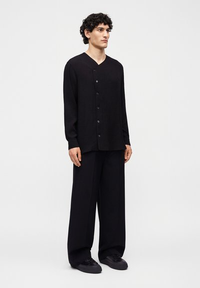 Νέος άνδρας στέκεται ελαφρώς στραμμένος προς τα δεξιά, φοράει φαρδιά μαύρο πουκάμισο με κουμπιά, φαρδιά μαύρα παντελόνια και μαύρα αθλητικά παπούτσια σε απλό φόντο.