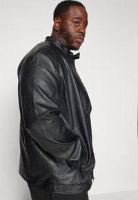 Chaqueta de cuero negra con un alto cuello, cierre de cremallera y bolsillos laterales. La textura suave y el corte relajado acentúan el diseño.