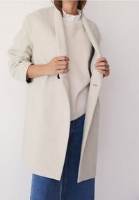 Manteau en laine gris clair à coupe décontractée et larges revers, superposé à un pull crème, associé à une jupe en denim bleu, mettant en valeur des textures douces.