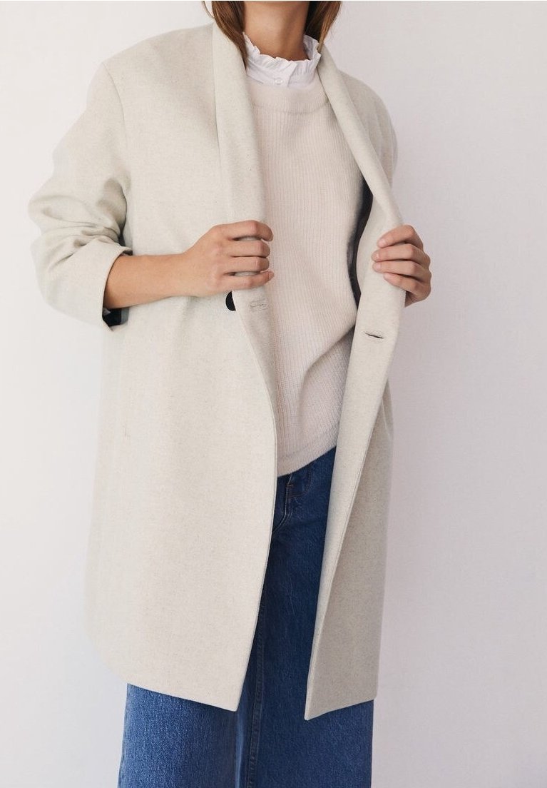 Manteau en laine gris clair à coupe décontractée et larges revers, superposé à un pull crème, associé à une jupe en denim bleu, mettant en valeur des textures douces.