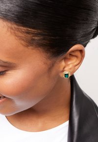 Pendientes de botón de oro con una piedra verde de corte esmeralda cuadrada, rodeada por un marco texturizado, que muestran elementos de diseño elegantes y modernos.