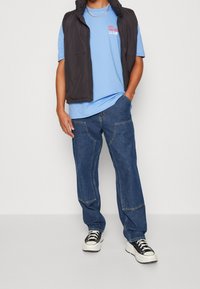 Svart lättviktsväst, blå t-shirt med röd text, blå löst sittande jeans med framfickor och svarta sneakers med vita sulor.