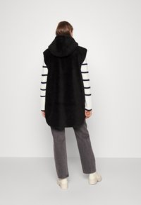 Czarna bezrękawnik z sztucznego futra z kapturem, w zestawieniu z białą i granatową koszulą w paski na długi rękaw, szarymi spodniami i beżowymi butami.