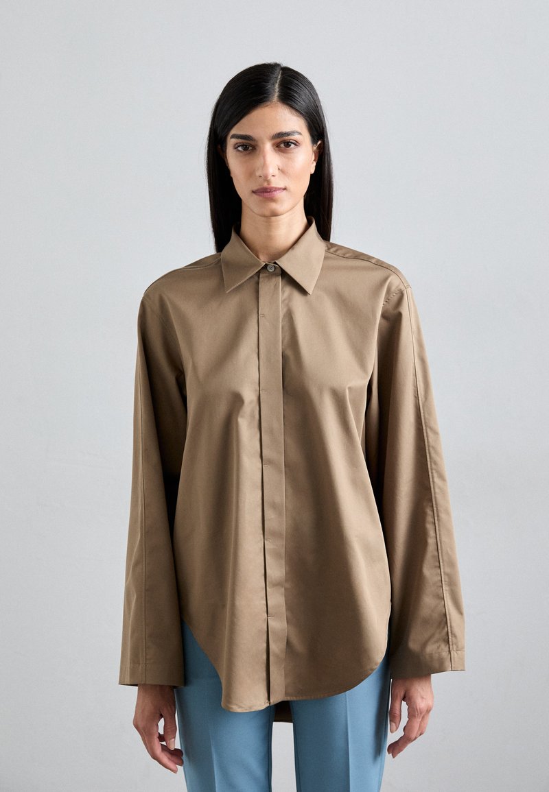 Brown, long-sleeve shirt with a pointed collar and button front, featuring a curved hem. Worn with blue, tailored pants. Smooth texture.